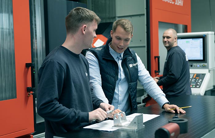 Two employees discuss a metal part while another employee operates the 5-axis milling machine